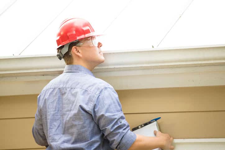 Repairman, building inspector, exterminator, engineer, insurance adjuster, or other blue collar worker has just begun his examination of a building/home's exterior walls, roof, and foundation. He wears a red hard hat and clear safety glasses and holds a his clipboard.